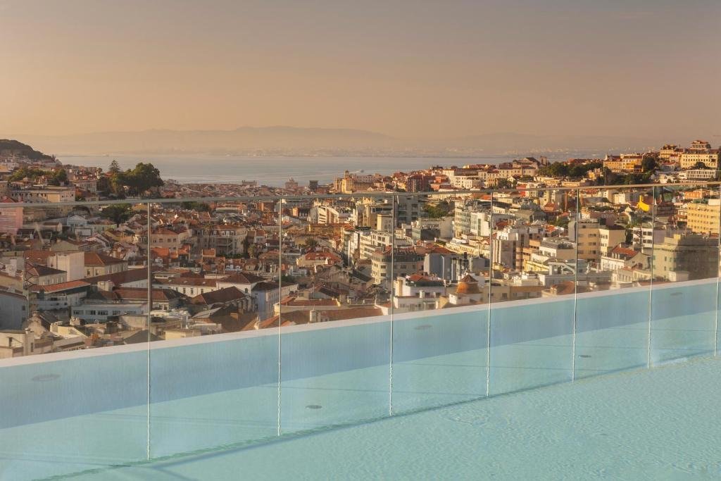 Rooftop pool with glass railing overlooking Lisbon cityscape and Tagus River under a warm, golden sunset.