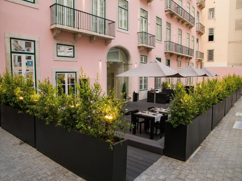 Pink building with balcony and outdoor patio seating, shaded by umbrellas, surrounded by greenery in large planters.