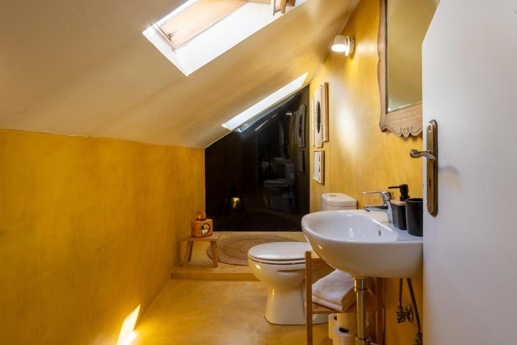 Cozy attic bathroom with slanted roof, yellow walls, skylight, sink, and toilet. Warm, inviting design and natural light.
