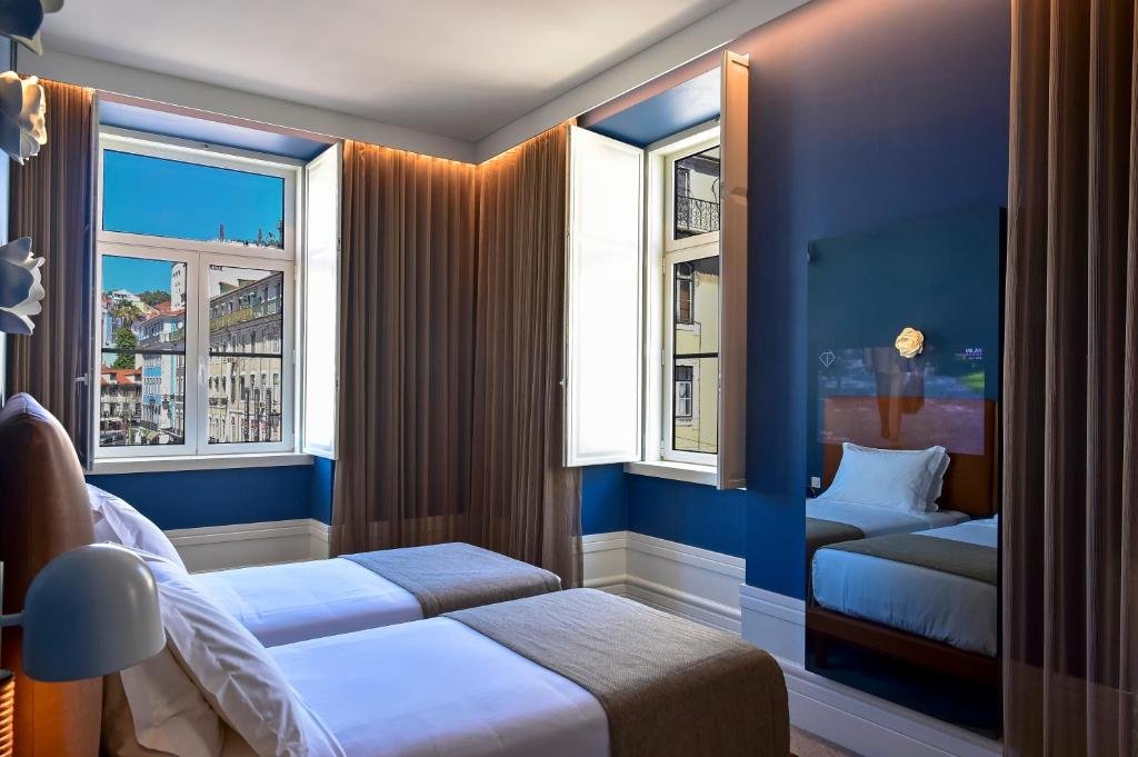 Modern hotel room with two beds, blue walls, and large windows offering a city view. Cozy and inviting atmosphere.
