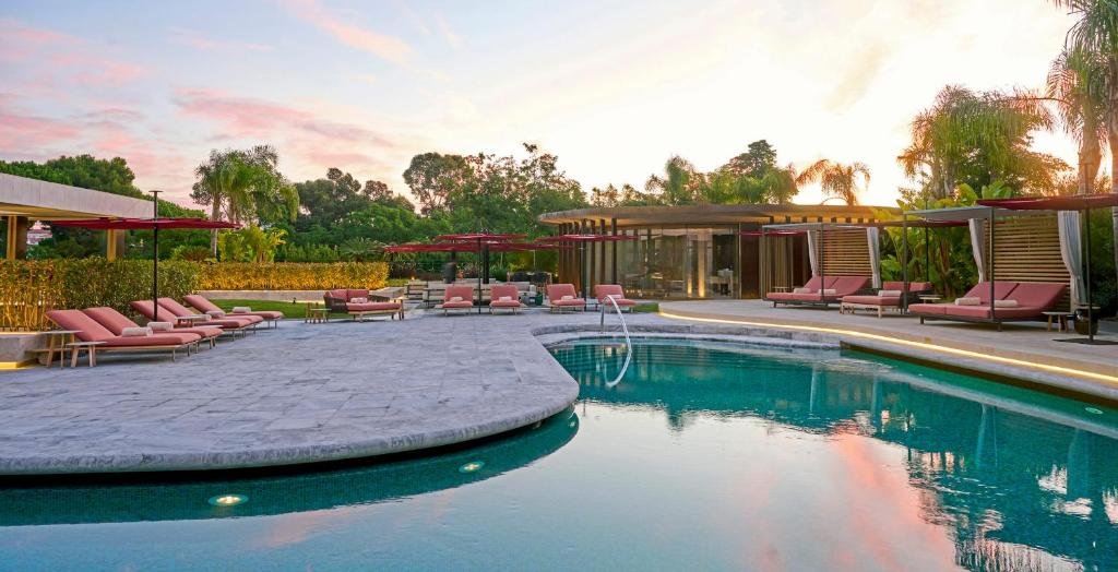 Luxury resort pool with pink lounge chairs and umbrellas at sunset, surrounded by lush greenery and palm trees.