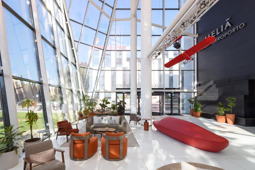 Modern hotel lobby with large windows, red airplane decor, chic furniture, and lush plants under a high ceiling.