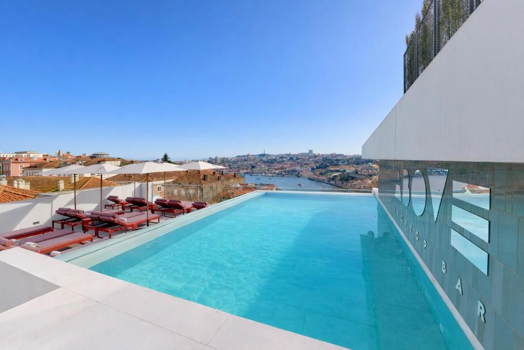 Rooftop pool with sun loungers and umbrellas overlooking cityscape and river under clear blue sky.