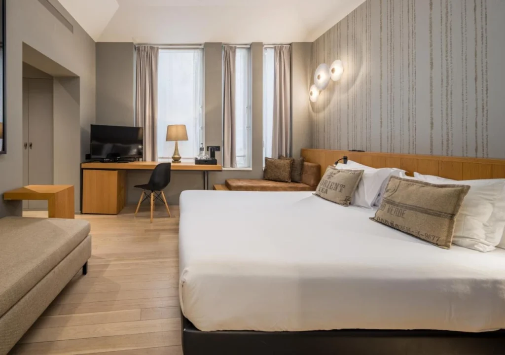 Modern hotel room with king bed, sofa, desk, flat-screen TV, large windows, and stylish lighting.