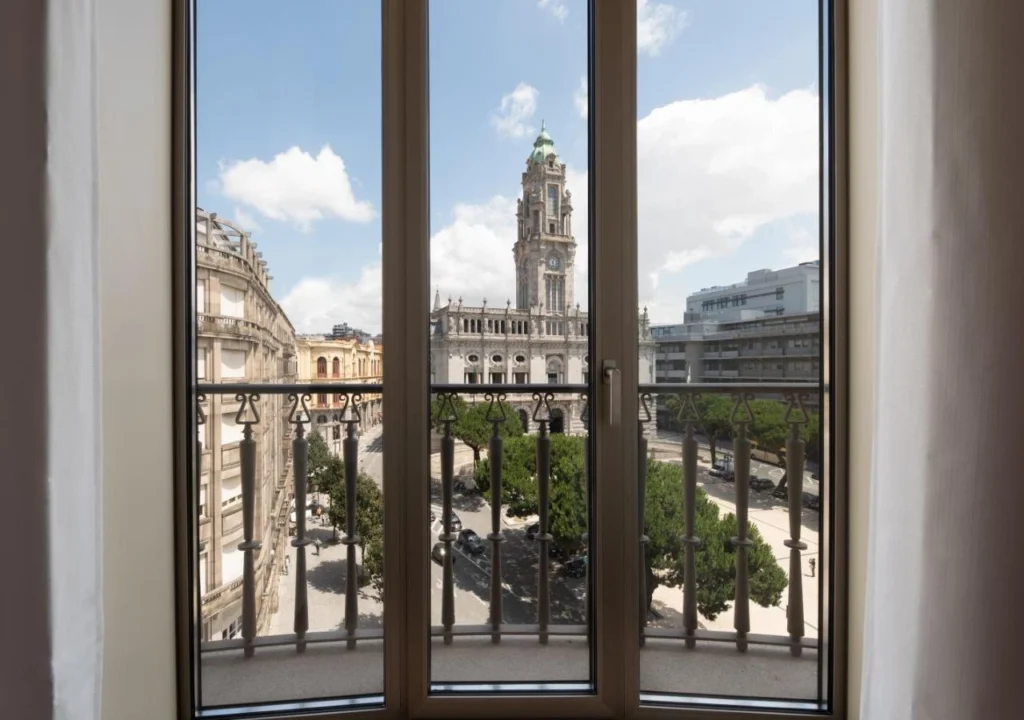 View of historic building through a window with balcony, featuring blue sky and cityscape.