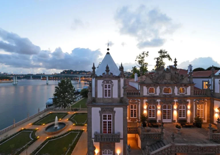 Elegant riverside mansion at dusk with illuminated gardens and fountain, overlooking a serene river and distant bridge.