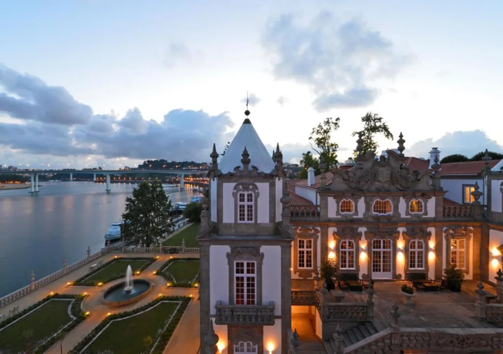 Historic mansion with lit facade overlooking a river and bridge at dusk, surrounded by lush gardens and a fountain.