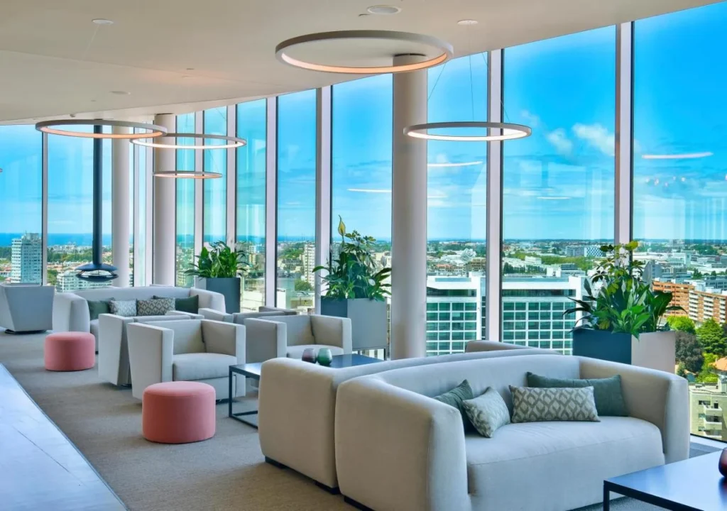 Modern lounge with sofas, large windows, city skyline view, and stylish lighting. Ideal for relaxation or meetings.