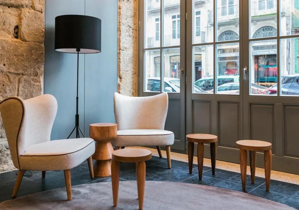 Modern lounge area with stylish chairs, wooden tables, and a floor lamp by large windows. Cozy interior design.