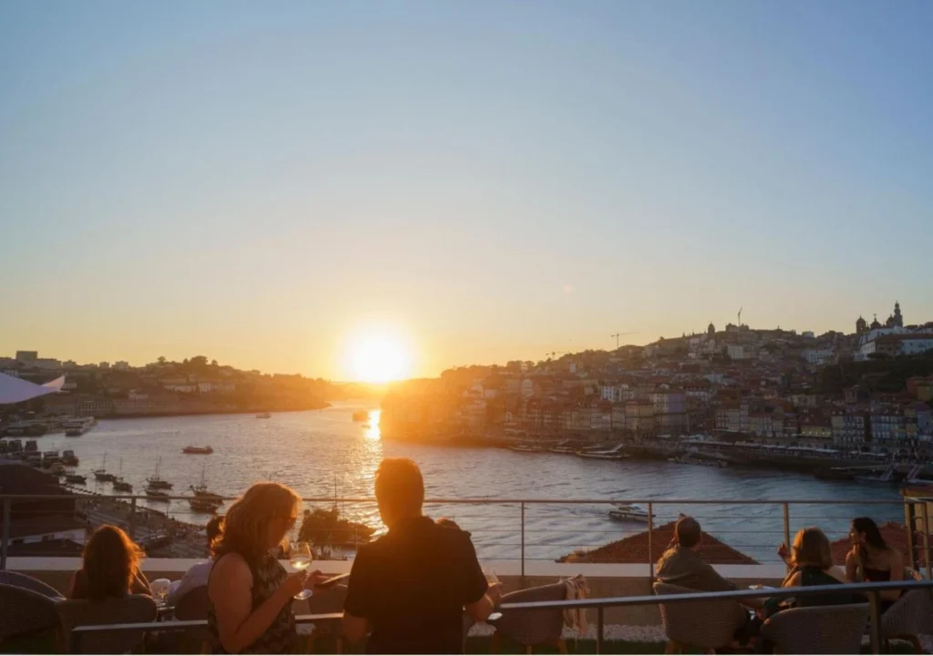 People enjoying a sunset view over a river and cityscape from a rooftop terrace, with boats visible on the water.
