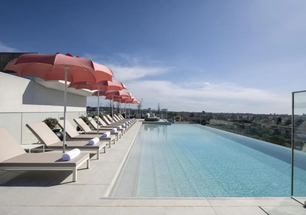Rooftop infinity pool with lounge chairs and umbrellas overlooking a cityscape under a clear blue sky.