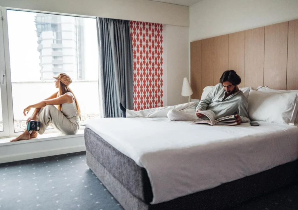 Couple relaxing in modern hotel room, woman gazing outside, man reading book on bed. Cozy and serene ambiance.