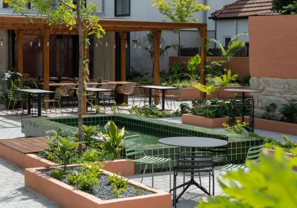 Modern outdoor patio with greenery, tables, and chairs under a wooden pergola, creating a fresh and relaxing ambiance.