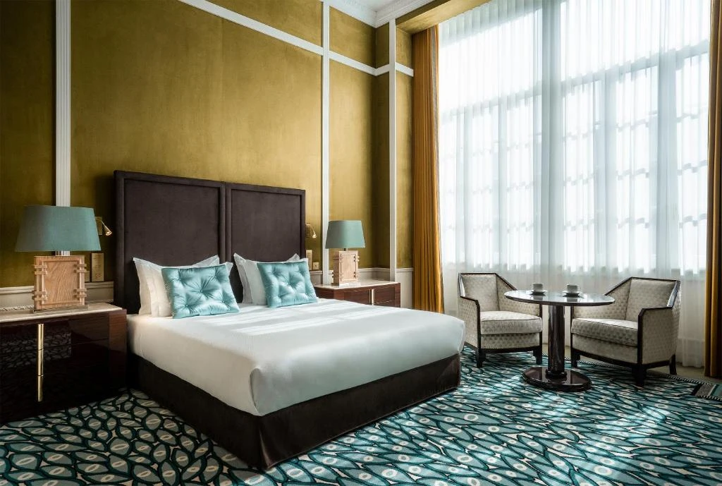 Elegant hotel room with a double bed, turquoise accents, and a seating area by a large window.
