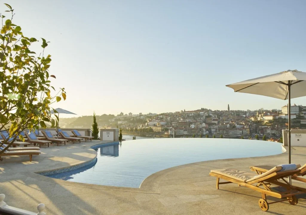 Infinity pool with city skyline view, surrounded by sun loungers and umbrellas under a clear blue sky.