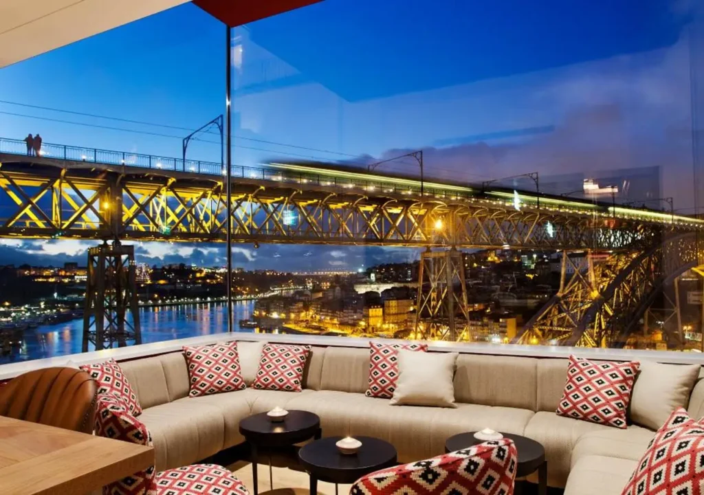 Cozy lounge with bridge and river view at dusk, featuring geometric cushions and ambient lighting.